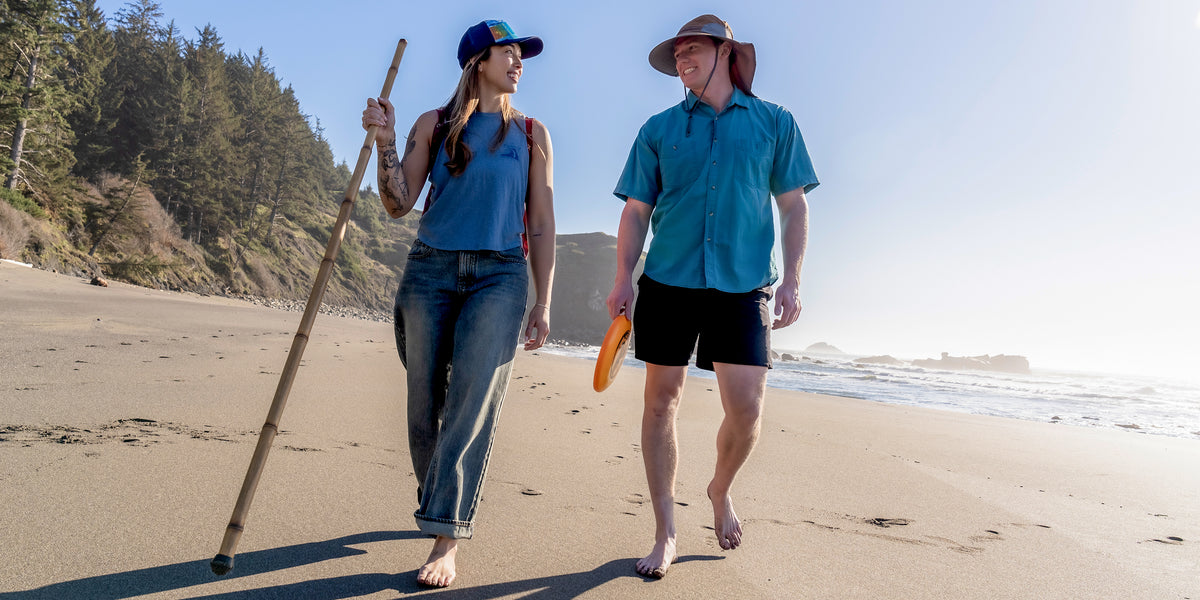 Two people walking on a beach with a stick and frisbee, surrounded by trees and water.